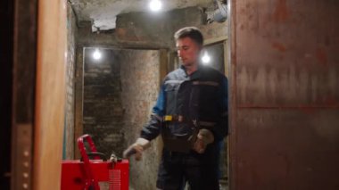 Profesyonel kaynakçı, kaynak makinesine gelir, elektrot tutacağı alır ve koruyucu maske takar. Genç işçi çalışma sürecine hazırlanıyor.