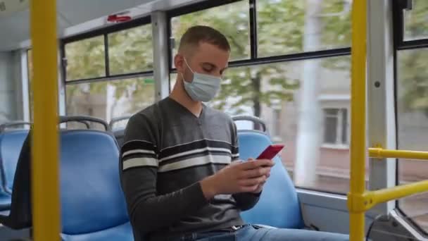 若い男性の乗客は 医療フェイスマスクでトロリーバスで旅行します カジュアルな服を着た男性はバスに座り スマートフォンを使用し コビド19から保護するために個人用保護装置の窓を見ます — ストック動画