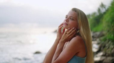 Cilt rutini, deniz kenarında ultraviyole koruma. Sarışın kadın yüzüne güneş kremi sürüyor, tropik sahillerde güneşlenmeyi seviyor. Güzellik, tatil, arka planda okyanus, rahatlık aktivitesi.