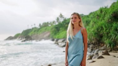 Huzurlu bayan okyanus esintisini, deniz kenarında rahat yürüyüşü sever. Mavi elbiseli zarif bir kadın kumlu sahilde geziniyor. Arka planda tropik dalgalar var. Kadın güzelliği, yaz modası, doğal kıyı manzarası..