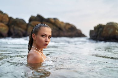 Denize dalmış, suya dayanıklı kozmetik ürünleri sergiliyor, sakin bakışlar atıyor. Su ortamında moda konusu, taşların arasında yüzme, dayanıklılık testi. Tatil atmosferi, yaz teması.