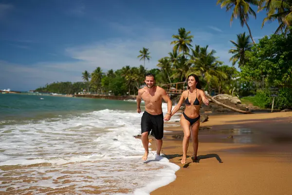 Muscular man and slender woman enjoy oceanfront run. Fit couple runs on sandy beach by the sea, surrounded by palm trees. Active lifestyle, couples workout, beach jogging in tropical paradise.