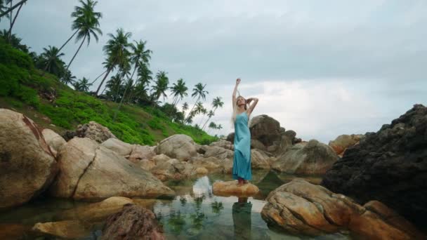 独行的女性享受自然之美 反映了在海滨的专注 身着蓝色长袍的优雅女士安详地站在巨石上 被平静的潮水包围 在海滨岩石和棕榈中 — 图库视频影像