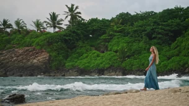 轻松自在的海风 舒缓的步行 在茂密的绿叶中的自然疗法 海浪爱抚沙滩 身着蓝色长袍的优雅女士漫步在宁静的热带海岸 — 图库视频影像