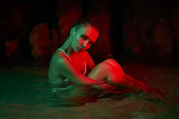 Female sits in lagoon at night, bathed in red green illumination. Artistic night photography, woman submerged in water. Beauty, inner self, wet model, creative colored light ambiance.