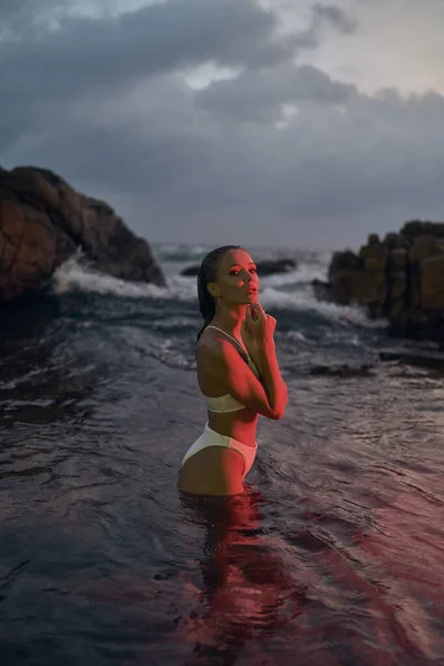 Beautiful woman relishing serene swim at night, sea bathed in glowing red hues. Lady in swimsuit finds peace in nocturnal ocean waters. Coastal vacation, tranquil retreat in natures aquatic setting.