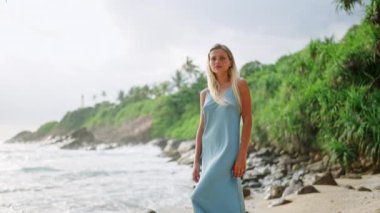 Mavi elbiseli sakin kadın deniz kenarında sakin bir tatilin tadını çıkarıyor. Zarif bir kadın kumlu sahilde gezer, tropik cennet ortamında. Cilt rutini, güneş koruması odak noktasında güzelliğin ortasında..