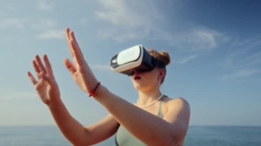 Sahil avatarı diyarının engin deneyimi, simülasyon gerçekliğiyle etkileşim, güneşli zemin, dalgalar kayalara çarpıyor. VR kulaklıklı kadın sanal deniz kenarını keşfediyor, havada jestler yapıyor..