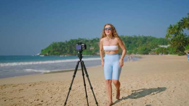 ダイナミックなダンスの動き ブロガーは海の背景に魅力的なコンテンツを作成します スタイリッシュなスポーツウェアの女性は トロピカルビーチで踊り ソーシャルメディアのための三脚でカメラでルーチンを記録 — ストック動画