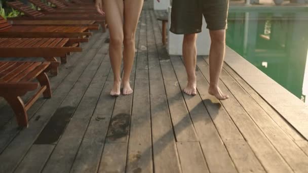 在热带旅馆的游泳池边 年轻的两只脚赤着脚走路 男友和女友的腿在热带度假酒店的木制甲板上手牵着手 在海滩俱乐部度过浪漫的假期 — 图库视频影像