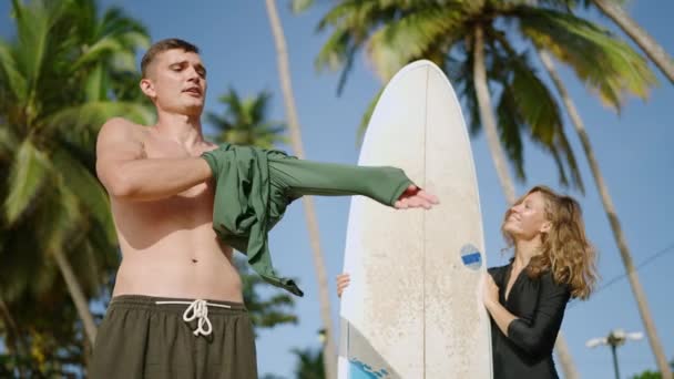 运动健美的年轻人穿上长袖皮疹护甲T恤 穿着泳衣的家伙穿着热带海滩上的椰子树 迷人的金发女人站在旁边 握着冲浪板 — 图库视频影像