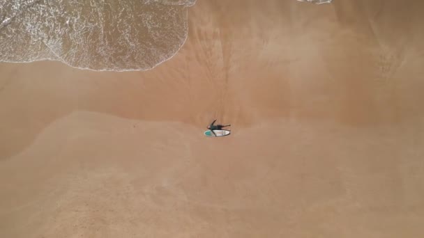 サーフボードで砂浜に横たわる水着の若者の空中トップビュー フィットサーファーの男は海の波の海サーフィンによって洗浄されるビーチの砂のサーフィンセッションの後でリラックスします ドローンが飛ぶ — ストック動画