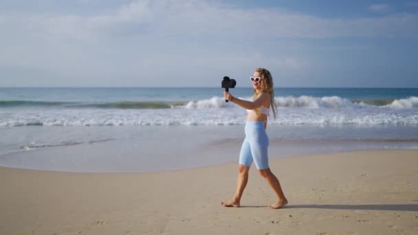 卷曲的头发的女人创造了满足 波浪在后面翻滚 蓝天在上面 女性旅行影响者记录在专业相机与无线Lav麦克风在沙滩上 个人旅行证件海滨旅行 — 图库视频影像