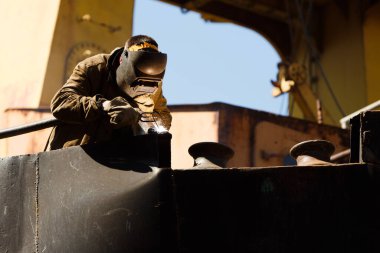 Kaynak maskeli bir zanaatkar kaynak yapar, ağır makinelerin arasındaki üretim sürecine odaklanır. Metal işleri uzmanlığı.
