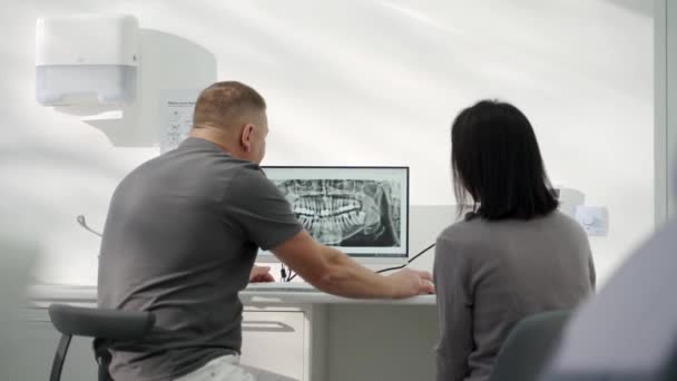 在牙科诊所 男牙医在电脑屏幕上向女病人展示口腔X光照片 正牙医生向妇女解释了屏幕上的X光牙齿 向牙科医生咨询 背景色 — 图库视频影像