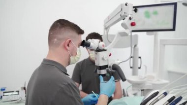 Modern stomatoloji kliniğinde diş mikroskobuyla çalışan erkek dişçi. Doktor kanal tedavisi için mikroskop kullanıyor. Stomatolog cerrahi operasyon gerçekleştirir. Kozmetik dişçiliği ve cerrahi.