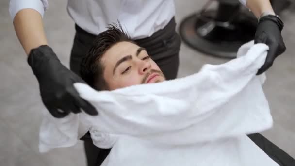 男客户放松 理发店洗胡子前的面部处理 理发师把热毛巾包在顾客的脸上 以便在沙龙里做剃须准备 传统的湿刮胡子概念 男用矿泉 — 图库视频影像