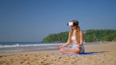 VR kulaklıklı kadın kumlu sahilde meditasyon yapıyor, denizin yanında sanal gerçeklik farkındalık seansı, teknoloji sağlığı trendi, kadın yogası yapıyor, VR zihinsel sağlık uygulaması sakin okyanus arkaplanı