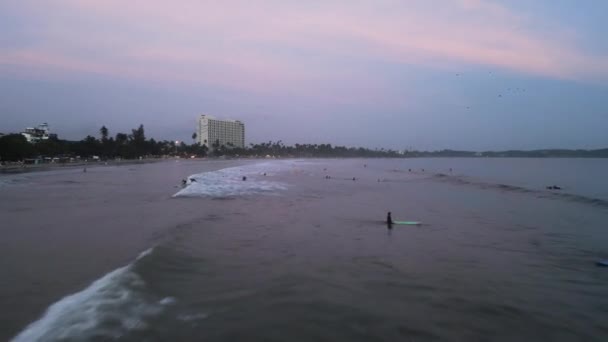 初出茅庐的冲浪者划着桨 像夕阳西下的天空 冲浪者黄昏时在拥挤的海里捕捉到海浪 空中景观显示了海岸线 海浪破浪 热带海滩上的冲浪课和酒店 — 图库视频影像