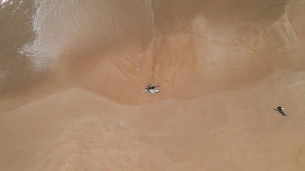 サーフボードで砂浜に横たわる水着の若者の空中トップビュー フィットサーファーの男は海の波の海サーフィンによって洗浄されるビーチの砂のサーフィンセッションの後でリラックスします ドローンが飛ぶ — ストック動画