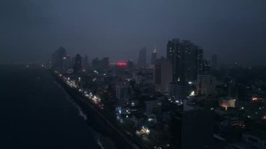 İnsansız hava aracı Sri Lanka başkentinin üzerinde uçuyor, sokakları aydınlatıyor, binalar vızıldayan şehir yaşamını özetliyor. Colombo 'nun gökyüzü gece görüşü, kıyı boyunca titreşen ışıklar, karanlık gökyüzünün altındaki şehir manzarası..