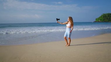 Sarışın etkisi seyahat videosu yaratır, kumlu sahilde yalınayak yürür. Modaya uygun kadın vlogger plajda dengeli kamerayla içeriği kaydediyor. Okyanus dalgalarının videografisi, tropikal manzara.
