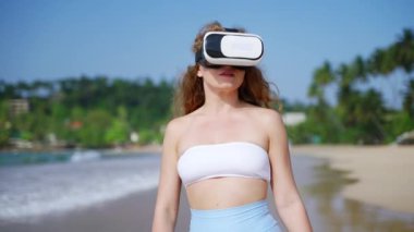 Meraklı kadın dijital dünyadan hoşlanıyor, modern eğlence teknolojisinden. Güneşli plajda VR kulaklıklı beyaz giysili bir kadın. Sanal gerçeklik, 3 boyutlu ortamlar. Yavaş çekim.