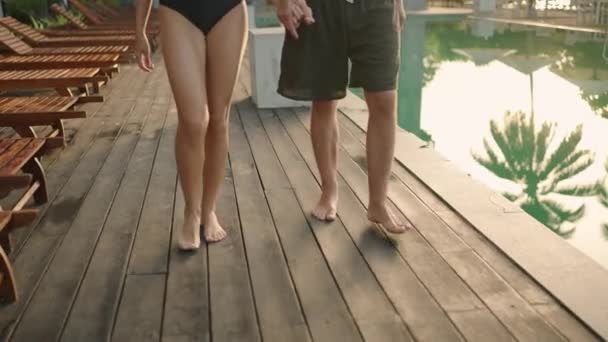 在热带旅馆的游泳池边 年轻的两只脚赤着脚走路 男友和女友的腿在热带度假酒店的木制甲板上手牵着手 在海滩俱乐部度过浪漫的假期 — 图库视频影像