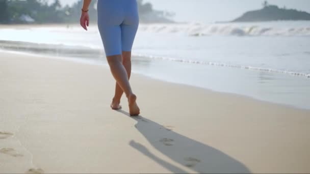 足迹在沙滩后面 健康撤退的感觉 日出或日落的感觉 海滨独行 赤脚人穿着蓝色短裤在沙滩上漫步 轻轻一挥 平静的海滨漫步 Slomo — 图库视频影像