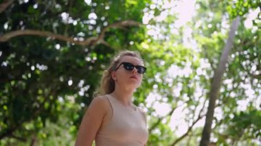 Bağımsız gezgin kadın yeşilliği keşfeder, başını dik tutar. Güneş gözlüklü aktif kadın yemyeşil ormanda doğa yürüyüşünden zevk alıyor, temiz hava soluyor. Sakin ağaçlarda tek başına macera, güneş ışığı filtresi..