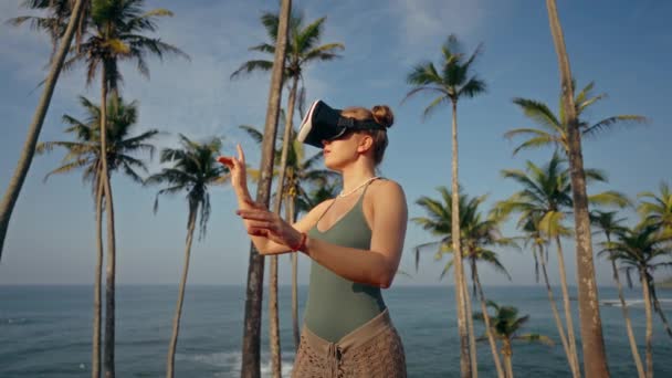 Vrヘッドセットの女性は トロピカルビーチで育ったバーチャルパームを探索し 没入型ゲームプレイでのジェスチャー 海洋による未来的なゲームに従事する屋外での拡張現実を体験します — ストック動画