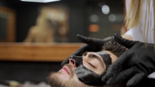 美容师在男人脸上平滑洁肤处理 理发师在美容院的男性顾客身上涂上黑色木炭面罩 用于皮肤护理 美容美发 男士化妆品 护肤例程 — 图库视频影像