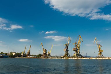 Denizcilik silueti, liman kenarı makineleri, ihracat-ithalat operasyonları, konteyner terminali, liman ekonomisi. Gemilere yük yükleyen vinçleri olan sanayi limanı, nakliye için ticaret merkezi, lojistik.