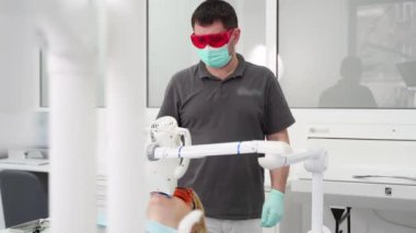 Profesyonel erkek dişçi, ultraviyole diş beyazlatmayı bitirdikten sonra hastaların koruyucu gözlüğünü çıkarıyor. Kırmızı gözlüklü konsantre doktor modern diş muayenehanesinde çalışıyor. Ağaran dişler.