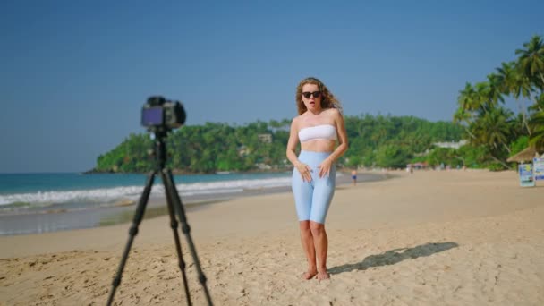 内容创建者为追随者制作卷轴 展示海洋中的运动 在热带海滩上 卷曲头发的女人穿着时髦的白色管状上衣和浅蓝色短裙 带着相机记录着跳舞的规律 — 图库视频影像