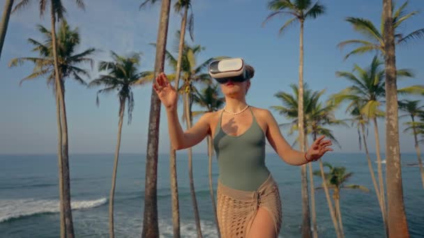 テクノロジー愛好家は 没入感のある現実 日没時の熱帯楽園でのジェスチャー 仮想旅行のトレンドコンセプトを体験します Vrヘッドセットの女性が海でヤシの木の間に仮想領域を探索する — ストック動画