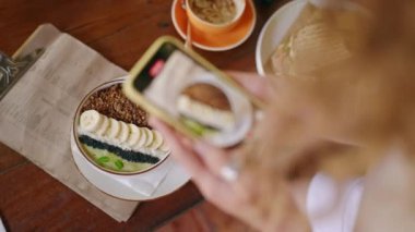Smoothie Bowl 'un fotoğraflarını çeken kadın eller, akıllı telefonlardan menüde. Lezzetli sandviç, masada kahve. Bali 'deki hippi kafesinde sağlıklı yemek fotoğrafları çeken bir kadın. Blogcu kahvaltı fotoğrafı çekiyor.