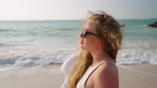 サングラスで歩くかわいい女の子 海岸で髪に触れる サーフィン 海岸で太陽浴をする若いモデル 海辺で夏休みを楽しんでいる魅力的な女性 クローズアップ — ストック動画