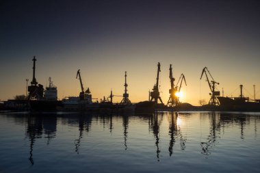 Endüstriyel deniz silueti. Yoğun limanda gündoğumu siluetleri kargo vinçleri. Ticaret gemileri kenetlendi, küresel lojistik, ihracat-ithalat operasyonları. Gündoğumu limanı, suyu yansıtıyor. Tedarik zinciri arkaplanı.