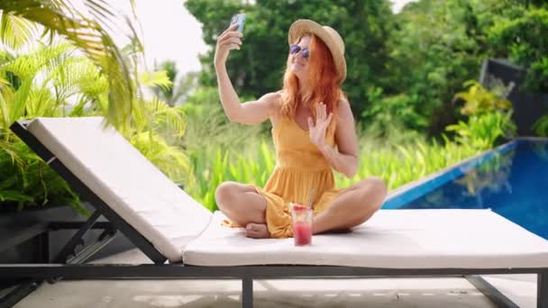 喝清爽的西瓜饮料 用手机拍自拍 穿着黄色衣服的红头发女人躺在蓝色的游泳池边 热带植物环抱着 在镜头前摇曳 享受着奢华的夏日 慢动作 — 图库视频影像