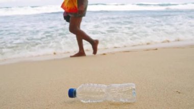 Kirlilik sorunu, insanların çöpleri görmezden gelerek deniz çevresi endişelerini vurgulamalarıyla ortaya çıkıyor. Çöpe atılmış plastik şişe kumlu sahilde uzanıyor ve yakınlarda dalgalar çarpıyor. Temizlik çalışmaları gerekiyor.