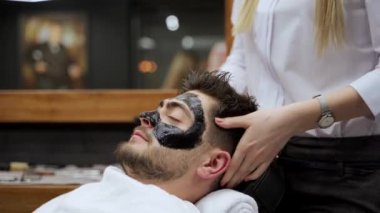 Adam berber koltuğunda derin temizlik sırasında rahatlıyor. Berber, siyah maskeyi müşterilerin yüzüne sürüyor. Cilt bakım rutininde. Güzellik uzmanı yüz kömürü ürünü.