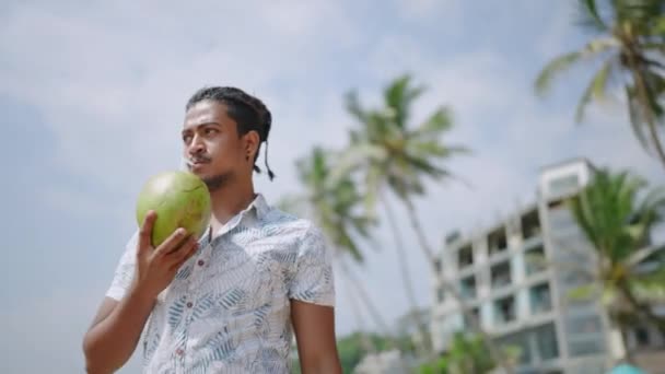 男性は緑のココを保持し 旅行のライフスタイルを楽しんでいます トロピカルビーチで新鮮なココナッツを吸い リラックス バジェットサバースのフルーツドリンク 暖かい気候レジャー — ストック動画