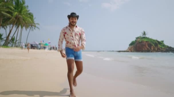 ケアフリーで表現力のある個人が休暇バイブ Lgbtqアイデンティティ ムーブメントを体現しています カウボーイのエネルギッシュな男 活気に満ちたシャツ デニムショートパンツは砂浜に沿ってダッシュし 熱帯の楽園に視聴者を招待 — ストック動画