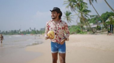 Kendine güvenen LGBTQ bireyi tropikal tatilin tadını çıkarıyor, neşe yayıyor, deniz kenarında özgürlük yayıyor. Kovboy şapkalı, çiçekli gömlekli, kumlu sahilde elinde hindistan cevizi olan bir adam. Palmiye ağaçlarının altında gündelik yürüyüş.