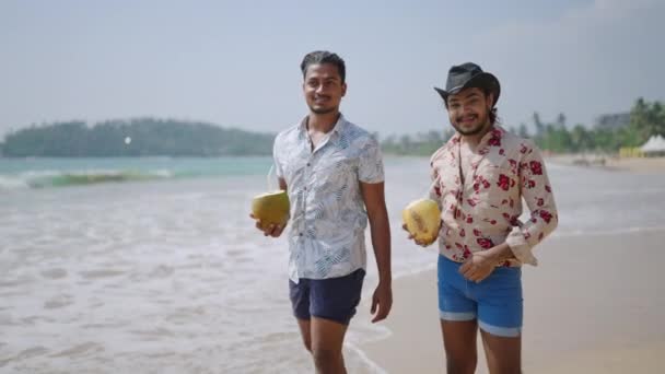 欢乐的热带假期 同性恋夫妇笑了 两个男人带着新鲜椰子在阳光灿烂的海滩上散步 海滨度假胜地的闲暇时间 花衬衫 牛仔帽反映节日时尚 — 图库视频影像