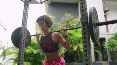 Spor giyim sporlarında atletik kadın, ton kasları. Çok ırklı, formda bir kadın dışarıda halterle çömeliyor. Dayanıklılık eğitimi, spor rutini motivasyonlu bireyler tarafından gösteriliyor. Yavaş çekim.