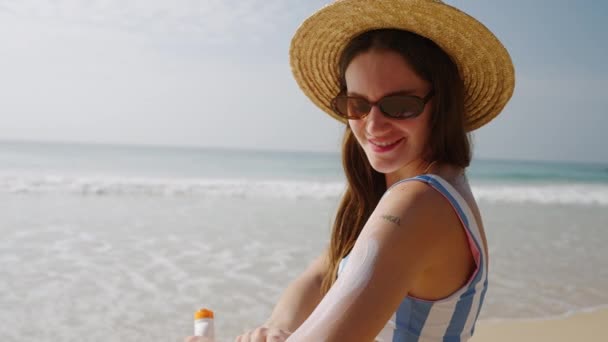 ハッピーな若い女性は海辺でスプーフサンローションを手に入れています わら帽子の美しい少女は紫外線から保護するために皮に防水日焼け止めを置きます 海でサンクリームを使用してビキニの女性の肖像画 — ストック動画
