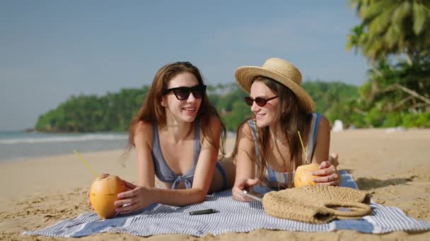 快乐的女孩在海滨晒日光浴 谈论着热带植物 美丽的女士躺在阳光下 在沙滩上浏览互联网 使用电话的年轻女性 在海滨用椰子鸡尾酒放松一下 — 图库视频影像