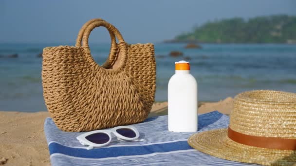わら帽子 サングラス タオルのトートバッグの隣に紫外線保護の日焼け止めボトル 夏の日の砂浜の海岸に配置されたビーチの必需品 日焼けに対するスキンケア 海辺でのレジャー — ストック動画